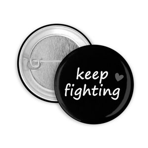 Könnte beinhalten: Schwarzer Knopf mit weißem Text, der "keep fighting" mit einem kleinen Herzsymbol lautet.