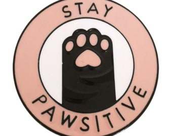 Può includere: Uno spillo in smalto rosa e bianco con un'impronta di zampa nera e il testo "STAY PAWSITIVE".