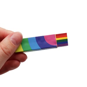 Könnte beinhalten: Eine hand hält eine regenbogenfarbene Schachtel mit einem Regenbogenmuster auf der Außenseite und einem Stapel regenbogenfarbener Gegenstände im Inneren. Die Schachtel ist rechteckig und hat ein farbenfrohes, geschwungenes Design.