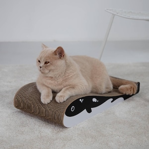 Può includere: Un gatto marrone chiaro riposa su un tiragraffi di cartone marrone. Il tiragraffi ha un design a balena in bianco e nero su un'estremità. Il gatto è sdraiato sul tiragraffi.