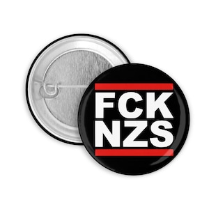 Può includere: Un bottone nero con una scritta bianca che dice "FCK NZS" con due linee rosse sopra e sotto il testo.