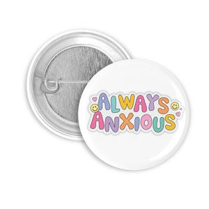 Könnte beinhalten: Zwei weiße, runde Ansteckbuttons. Einer hat eine Nadel auf der Rückseite, der andere ein farbenfrohes Design. Das Design zeigt "ALWAYS ANXIOUS" in Buchstaben mit Herzen und Smileys.