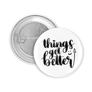 Könnte beinhalten: Weißer Knopf mit schwarzem Text, der "things get better" mit einem nach oben zeigenden Pfeil und einem Smiley sagt.