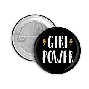 Könnte beinhalten: Ein schwarzer Ansteckbutton mit den weißen Worten "GIRL POWER", akzentuiert mit goldenen Blitzsymbolen. Die Rückseite des Buttons ist silberfarben mit einer Nadel.