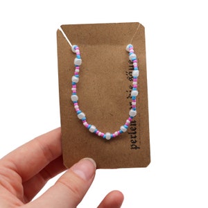 Könnte beinhalten: Ein Perlenarmband mit weißen, rosa und blauen Perlen auf einer braunen Karte mit dem aufgedruckten Text "perlen-liebe".