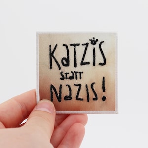 Könnte beinhalten: Ein quadratischer Stoffaufnäher mit einem beigen und braunen Farbverlauf. Der Aufnäher zeigt den schwarzen gestickten Text KATZIS STATT NAZIS! mit einem kleinen Kronensymbol über dem I in Katzis und einem weißen, gestickten Rand.