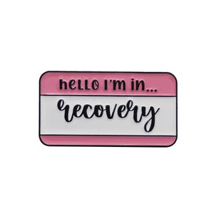 Könnte beinhalten: Rosa-weiße Emaille-Anstecknadel mit dem Text "hello i'm in... recovery" in schwarzer Schrift.
