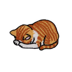Pode incluir: Remendo bordado de um gato malhado laranja dormindo com listras marrons e detalhes brancos. O gato está enroscado, com os olhos fechados e as patas dobradas. O remendo tem uma borda preta.