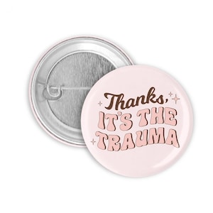 Könnte beinhalten: Ein rosa Knopf mit braunem Text, der "Danke, es ist das Trauma" lautet. Der Knopf hat eine silberne Rückseite.