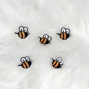 May include: Five embroidered bee patches. Each patch features a black and orange striped body, white wings, and a black outline. The patches are small, measuring approximately 1 inch in length, and are set against a white, fluffy background.