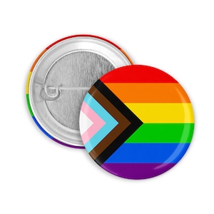 May include: Two round pin badges. One badge features a rainbow design with a black, brown, white, pink, and light blue chevron. The other badge has a silver metal back and a pin.