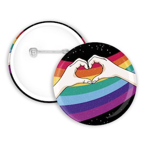 Op de afbeelding: Een regenboog-pride-knop met een zwarte achtergrond en witte sterren. Twee handen vormen een hart boven de regenboog.