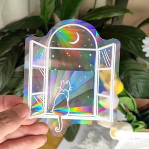 Könnte beinhalten: Holografischer Aufkleber mit weißer Umrisszeichnung einer Katze, die aus einem Fenster einen Halbmond und Sterne betrachtet. Das Fenster ist offen, und der Hintergrund hat ein regenbogenfarbenes Sonnenstrahlendesign. Der Aufkleber wird von einer Hand gehalten.