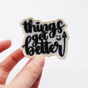 Könnte beinhalten: Weißer gestickter Aufnäher mit schwarzer Aufschrift "things get better" mit einem Smiley im "get" Teil des Textes.