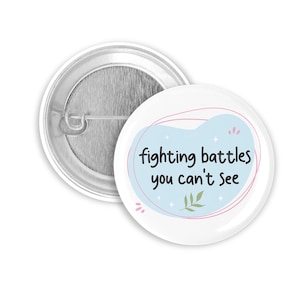 Könnte beinhalten: Ein weißer Ansteckbutton mit dem Text "fighting battles you can't see" in Schwarz. Der Text befindet sich in einer hellblauen Form mit rosa und grünen Akzenten. Der Button hat eine silberne Metallrückseite.