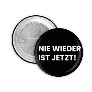 Könnte beinhalten: Ein schwarzer Button mit dem weißen Text "NIE WIEDER IST JETZT!". Der Button hat eine Anstecknadel und einen silbernen Metallrand. Der Button hat eine runde Form.
