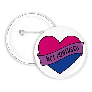 Könnte beinhalten: Ein weißer Knopf mit einem pinkfarbenen, violetten und blauen Herzen mit einem violetten Banner, auf dem "NOT CONFUSED" steht.