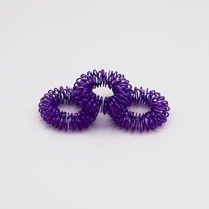 Könnte beinhalten: Drei violette Fingermassageringe sind vor einem weißen Hintergrund angeordnet. Jeder Ring besteht aus gewickeltem Metall und ist für die Verwendung an den Fingern bestimmt. Die Ringe haben einen leuchtenden Lilaton.