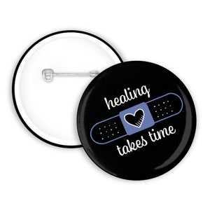 Könnte beinhalten: Schwarzer Knopf mit einem weißen Verband mit einem Herzen und dem Text "healing takes time".