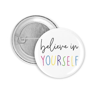Può includere: Un bottone bianco con una scritta in corsivo nero "believe in" e la parola "YOURSELF" in colori arcobaleno.
