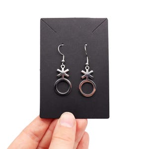 Könnte beinhalten: Ein Paar silberfarbene Ohrringe mit einem Gender-Symbol-Design. Jeder Ohrring hat einen Kreis mit einem Kreuz darüber und einen Haken. Die Ohrringe werden auf einer schwarzen Karte präsentiert.
