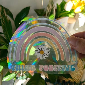 Könnte beinhalten: Ein holografischer Aufkleber mit einem Regenbogen-Design in Pastellfarben und einer Gänseblümchen-Blume in der Mitte. Der Aufkleber trägt den Schriftzug "THINK POSITIVE" in einer bunten Retro-Schrift. Der Aufkleber wird vor einem Hintergrund aus grünen Pflanzen gehalten.