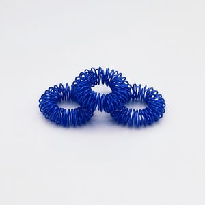 Könnte beinhalten: Drei blaue Fingermassageringe. Jeder Ring besteht aus aufgerolltem Metall und ist so konzipiert, dass er an den Fingern auf und ab gerollt werden kann. Die Ringe haben eine leuchtend blaue Farbe und sind auf weißem Hintergrund angeordnet.