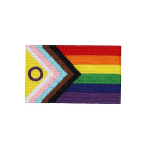 Può includere: Una bandiera arcobaleno del pride con un cerchio viola e un centro giallo, che rappresenta l'inclusione intersessuale, è cucita su un triangolo nero, marrone, azzurro, rosa e giallo. La bandiera è fatta di tessuto e ha una trama tessuta.