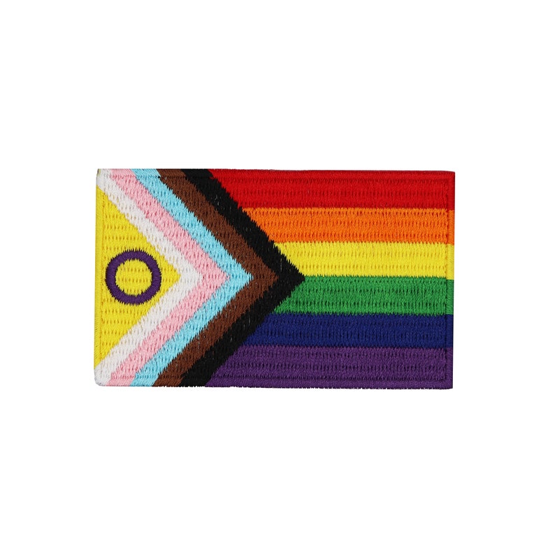 May include: A rainbow pride flag with a purple circle and a yellow center, representing intersex inclusion, is sewn onto a black, brown, light blue, pink, and yellow triangle. The flag is made of fabric and has a woven texture.