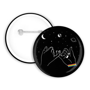 Könnte beinhalten: Ein schwarzer Button mit einer weißen Umrandung von zwei Händen, die ein Versprechen mit dem kleinen Finger geben, vor einem Sternenhintergrund mit Mond, Planet und Sternen. Ein Regenbogen-Pride-Flag ist auf einem Handgelenk.