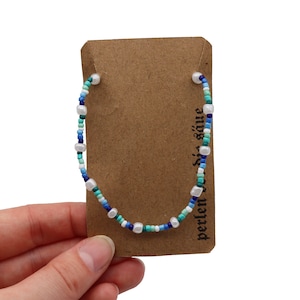 Könnte beinhalten: Ein Perlenarmband mit blauen, türkisfarbenen und weißen Perlen. Das Armband ist auf einer transparenten Schnur aufgefädelt und wird auf einer braunen Karte präsentiert. Die Karte hat Text in einer Fremdsprache.