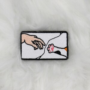Könnte beinhalten: Gestickter Aufnäher mit einer Parodie von Michelangelos 'Erschaffung Adams', bei der eine menschliche Hand auf eine Katzenpfote zeigt. Der Aufnäher hat einen schwarzen Rand und einen weißen Hintergrund. Die Katzenpfote hat rosa Ballen und orangefarbene Akzente.