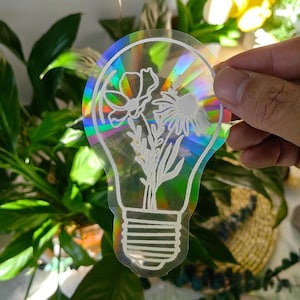 Könnte beinhalten: Ein klarer, holografischer Aufkleber in Form einer Glühbirne mit einem weißen Blumendesign. Der Aufkleber hat einen Regenbogeneffekt, wobei sich das Licht durch das transparente Material bricht. Das Blumendesign umfasst verschiedene Blumen und Blätter.