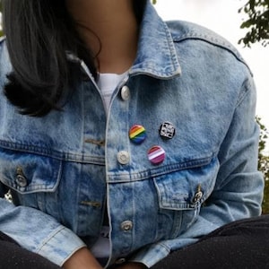 Könnte beinhalten: Eine Person trägt eine blaue Jeansjacke mit drei Anstecknadeln. Die Anstecknadeln sind eine Regenbogenflagge, ein pink-weiß gestreiftes Herz und ein schwarz-weißes Quadrat mit dem Text "Schütze Trans-Kinder".