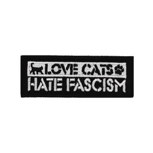 Könnte beinhalten: Schwarzer rechteckiger Aufnäher mit weiß gesticktem Text und einer Katzen-Silhouette. Der Text lautet "LOVE CATS HATE FASCISM" mit einer Pfotenabdruck-Grafik. Der Aufnäher ist wahrscheinlich für Kleidung oder Accessoires.