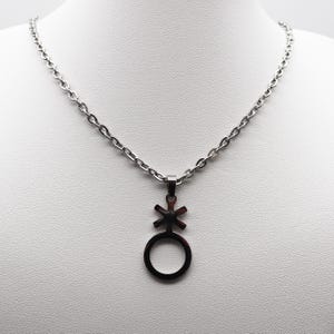 Puede incluir: Un collar de cadena de plata con un colgante que presenta el símbolo de género neutro. El símbolo es un círculo con una cruz en su interior.
