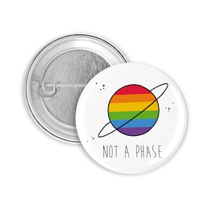 Könnte beinhalten: Ein weißer Knopf mit einem Regenbogenplaneten und dem Text "NOT A PHASE" in Schwarz.