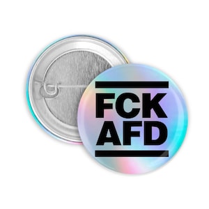 Könnte beinhalten: Ein runder Knopf mit holografischem, schimmerndem Hintergrund. Der Knopf hat schwarzen Text, der "FCK AFD" lautet, mit zwei schwarzen Linien darüber und darunter.