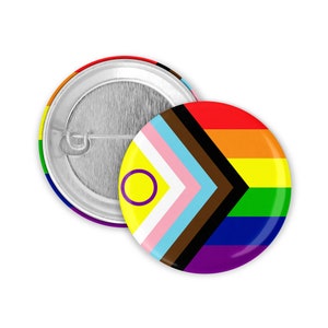 May include: Two round pin badges. One badge features a rainbow design, the other has a yellow circle with a purple outline, a pink and light blue chevron, and brown and black stripes. The badges are on a white background.