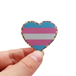 Könnte beinhalten: Ein herzförmiger Anstecker mit dem Design der Transgender-Pride-Flagge. Die Flagge hat horizontale Streifen in Hellblau, Rosa und Weiß. Der Anstecker hat einen glitzernden Goldrand und wird von einer Person gehalten.