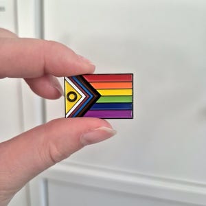 Op de afbeelding: Een regenboogvlagpin met een gele cirkel en een zwarte driehoek in de linkerbovenhoek. De vlag heeft strepen in rood, oranje, geel, groen, blauw, indigo en violet.