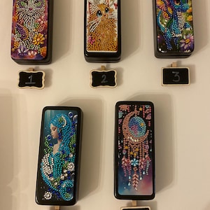 May include: Five small black boxes with different colorful images on the front. The images are of a cat, a dragon, a woman, a moon, and a dream catcher. Each box has a small black sign with a number on it, from 1 to 5.