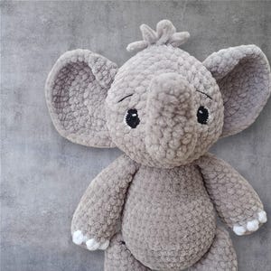 May include: A grey crocheted elephant toy with large ears and a trunk. The elephant has black and white embroidered eyes and white accents on its feet. The toy is made of a soft, textured material.