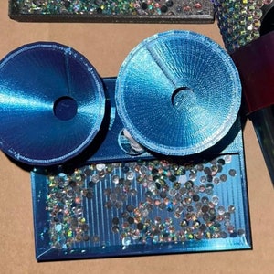 Op de afbeelding: Een blauwe dienblad met een glinsterend, regenboogkleurig oppervlak. Twee blauwe, ronde decoratieve stukken met een gat in het midden zijn bevestigd aan het dienblad.