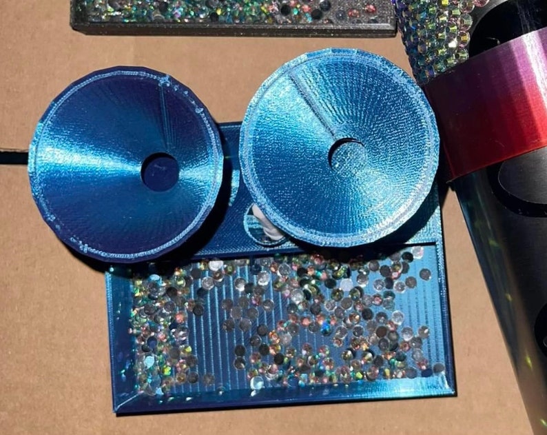 Op de afbeelding: Een blauwe dienblad met een glinsterend, regenboogkleurig oppervlak. Twee blauwe, ronde decoratieve stukken met een gat in het midden zijn bevestigd aan het dienblad.