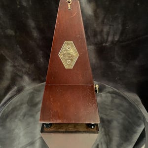 May include: A vintage wooden metronome with a pyramid shape. The metronome is a dark brown color with a brass-colored emblem on the front. The metronome sits on a clear glass surface.