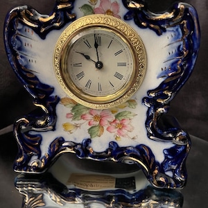 May include: An antique porcelain clock with a white face, gold-tone bezel, and Roman numerals. The clock's frame is white with blue and gold accents, and floral designs. The clock face reads "The Ansonia Clock Co. New York, United States of America."