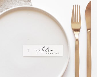 May include: White place setting with a gold fork and knife. A white card with the name "Andrew Raymond" is placed on the plate.