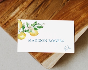 May include: White place card with a watercolor illustration of lemons and blossoms. The text "MADISON ROGERS" is printed in blue text. The card is on a wooden surface.