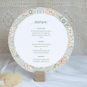 May include: A circular menu card featuring a decorative border of sage green, terracotta, and white geometric patterns. The center displays the text menu, including appetizer, entrée, and dessert sections, held upright by a small wooden block stand.